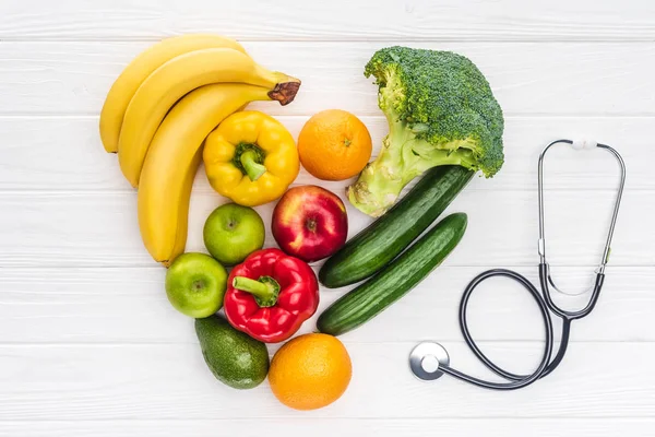 Vista superior del símbolo del corazón hecho de frutas y verduras frescas y estetoscopio en la superficie de madera - foto de stock