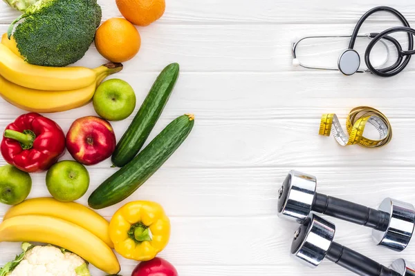 Vista dall'alto di frutta e verdura fresca, manubri, stetoscopio e metro su tavolo in legno — Foto stock