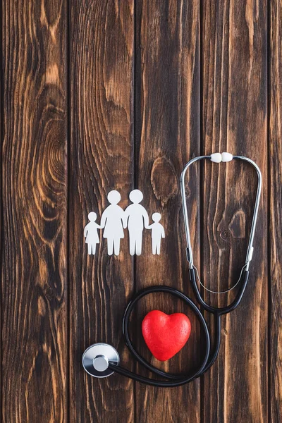 Draufsicht auf weiße Familienfiguren, die sich an Händen halten, rotes Herzsymbol und Stethoskop auf Holztisch — Stockfoto