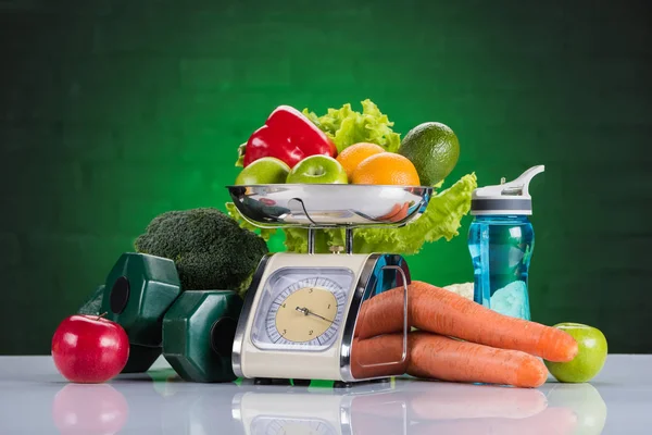 Close-up view of fresh fruits and vegetables on scales, dumbbells and bottle of water on green — Stock Photo