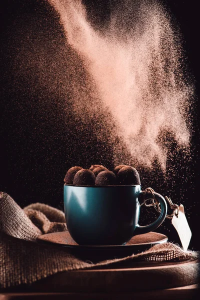 Closeup image of grated chocolate falling on truffles in cup of coffee on sackcloth and cutting board on black background — Stock Photo