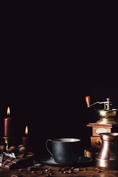 Closeup shot of cup of coffee at wooden table with chocolate, truffles, coffee grains, candles and turk on black background — Stock Photo