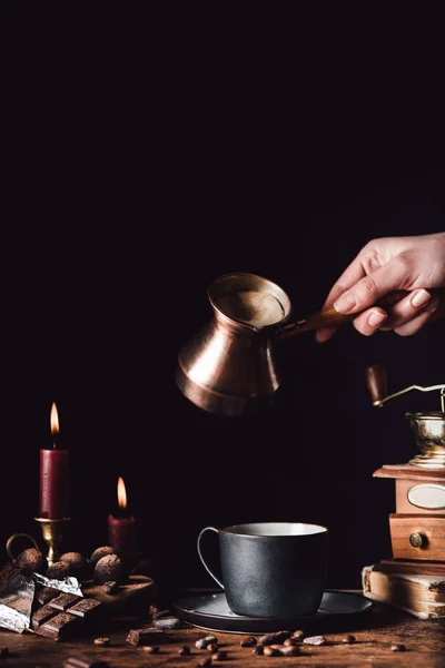 Partial view of woman pouring coffee from turk into cup at table with chocolate, truffles, candles and coffee grains on black background — Stock Photo