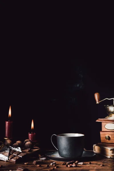 Closeup shot of cup of coffee at wooden table with chocolate, truffles, coffee grains and candles  on black background — Stock Photo