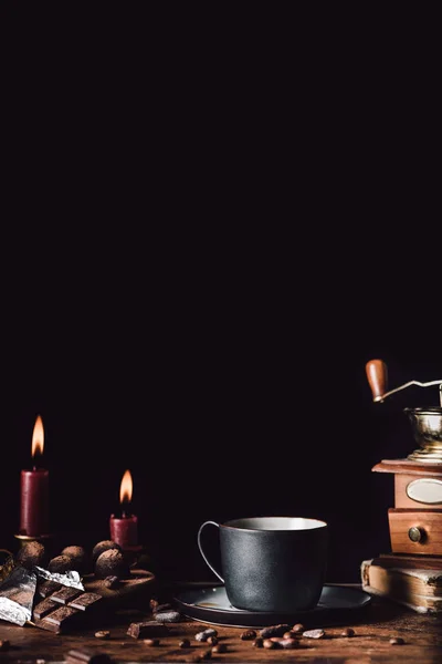 Close up image of cup of coffee at wooden table with chocolate, truffles,coffee grains and candles on black background — Stock Photo