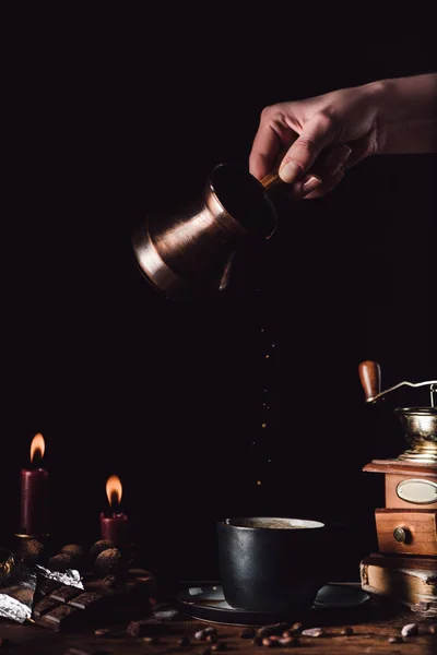 Partial view of woman pouring coffee from turk into cup at table with chocolate, truffles, candles and coffee grains on black background — Stock Photo