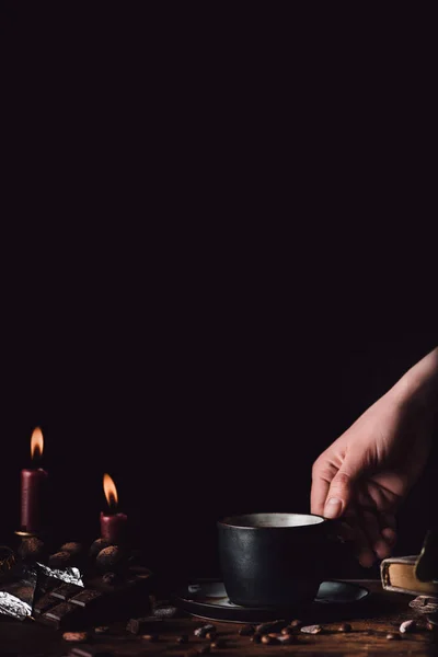 Cropped image of woman holding cup of coffee at wooden table with chocolate, truffles, coffee grains, candles and book on black background — Stock Photo