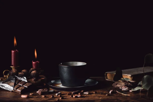 Nahaufnahme einer Tasse Kaffee am Holztisch mit Schokolade, Trüffeln, Kaffeekörnern, Kerzen und Buch auf schwarzem Hintergrund — Stockfoto