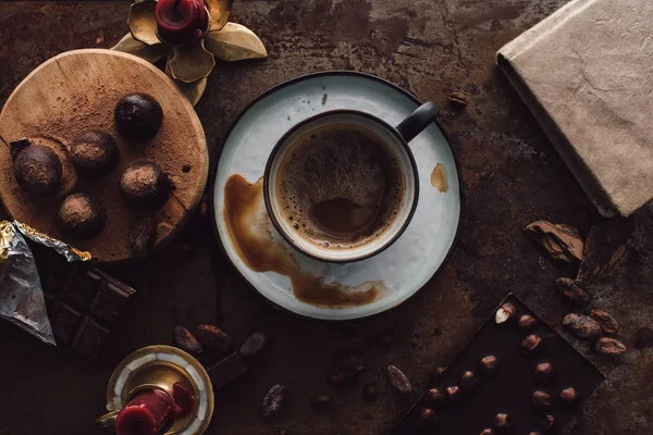 Високий вид чашки кави, трюфелі на дерев'яному скибочці, свічки, какао-боби, книги та два шоколадні батончики на сільському столі — стокове фото