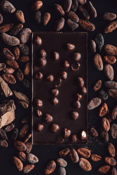 Vista superior da barra de chocolate com avelãs cercadas por grãos de cacau na superfície escura — Fotografia de Stock