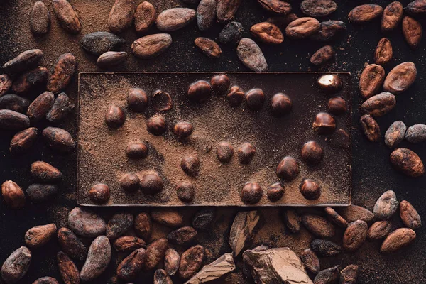 Vista sopraelevata di tavoletta di cioccolato con nocciole circondata da fave di cacao ricoperte di cioccolato grattugiato — Foto stock
