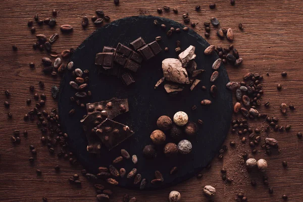 Elevated view of cutting board with various types of chocolate pieces and truffles surrounded by cocoa beans, coffee grains and nutmegs on wooden table — Stock Photo