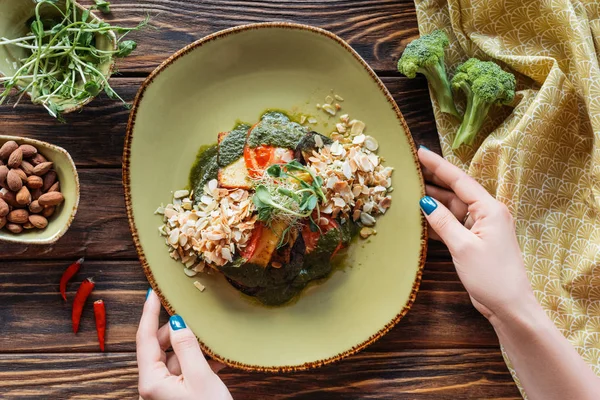 Vista parziale di donna che tiene piatto di insalata vegetariana servita con mandorle grattugiate e germogli su tavolo in legno — Foto stock