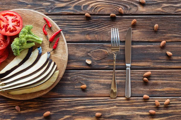 Vue de dessus de l'aubergine fraîche coupée, tomates, brocoli et piments sur planche à découper sur plateau en bois avec couverts et amandes autour — Photo de stock