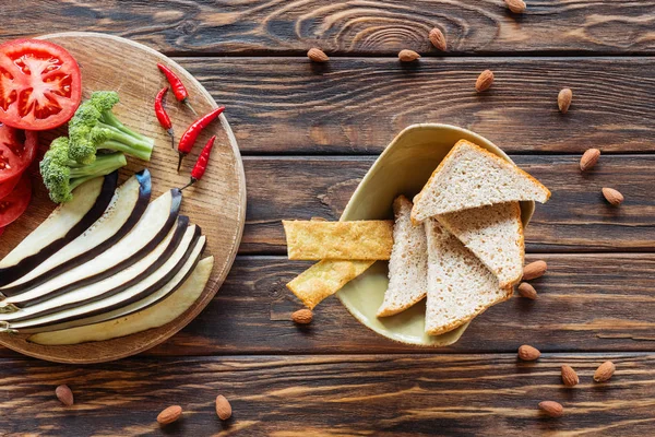 Top view of fresh cut eggplant, tomatoes, broccoli and chili peppers on cutting board on wooden tabletop with bowl on bread — Stock Photo