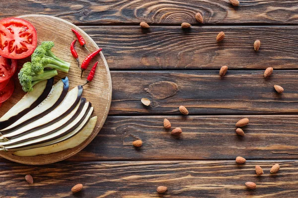 Top view of fresh cut eggplant, tomatoes, broccoli and chili peppers on cutting board on wooden tabletop with almonds around — Stock Photo