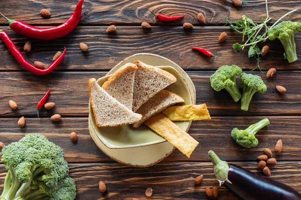 Flat lay with pieces of bread in bowl, almonds, chili peppers and fresh vegetables around on wooden tabletop — Stock Photo