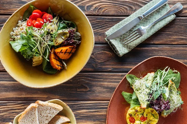 Flat lay com saladas vegetarianas servidas em tigelas e talheres em mesa de madeira — Fotografia de Stock