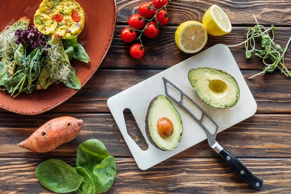 Deitado plano com salada vegetariana, abacate corte na tábua de corte, faca e legumes frescos em torno de mesa de madeira — Fotografia de Stock