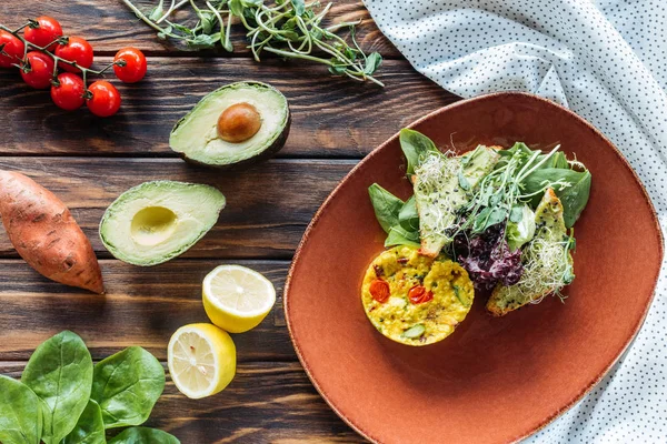 Flat lay com salada vegetariana servida em prato e ingredientes frescos dispostos em torno de mesa de madeira — Fotografia de Stock