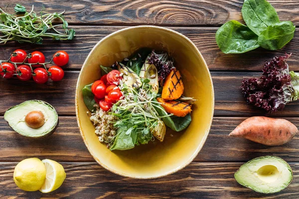 Acostado plano con ensalada vegetariana con verduras a la parrilla, brotes, tomates cherry en un tazón e ingredientes frescos dispuestos alrededor en la mesa de madera - foto de stock