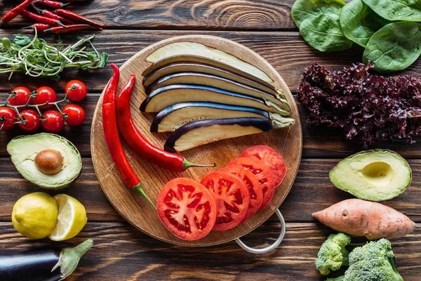 Flat lay with cut eggplant, tomatoes and chili peppers on cutting board with fresh vegetables around on wooden surface — Stock Photo