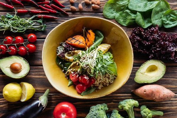 Flat lay com salada vegetariana com legumes grelhados, brotos, tomates cereja na tigela e ingredientes frescos dispostos em torno de mesa de madeira — Fotografia de Stock