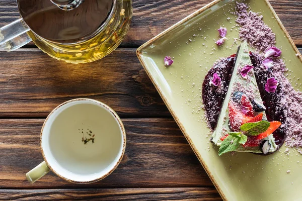 Draufsicht auf arrangiertes vegetarisches Dessert mit Beeren auf Teller, leerer Tasse und Teekanne auf Holzoberfläche — Stockfoto