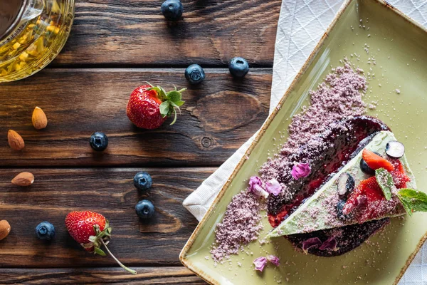 Vista superior del postre vegetariano arreglado con bayas en plato, almendras y tetera en superficie de madera - foto de stock