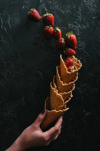 Female hand holding waffle cones with summer strawberries on dark background — Stock Photo