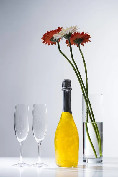 Vista de cerca de las copas vacías, botella de champán y ramo de flores de gerberas sobre fondo gris - foto de stock