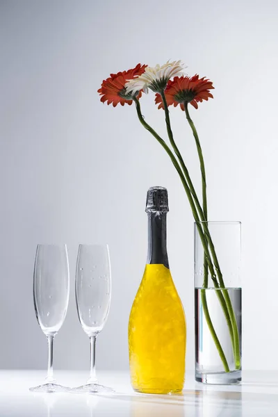 Close up view of empty glasses, bottle of champagne and bouquet of gerbera flowers on grey backdrop — Stock Photo