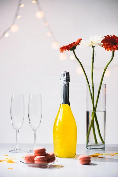 Vista de cerca de macarrones, copas vacías, botella de champán y ramo de flores de gerberas sobre fondo gris - foto de stock