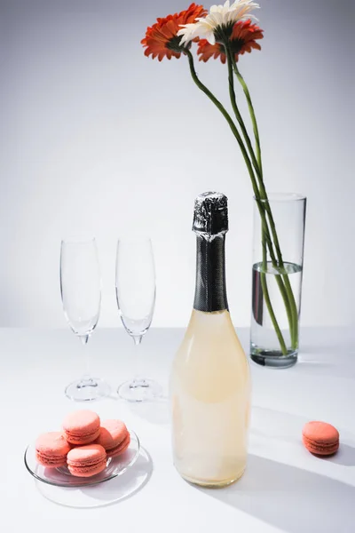 Vista de cerca de macarrones, copas vacías, botella de champán y ramo de flores de gerberas sobre fondo gris - foto de stock