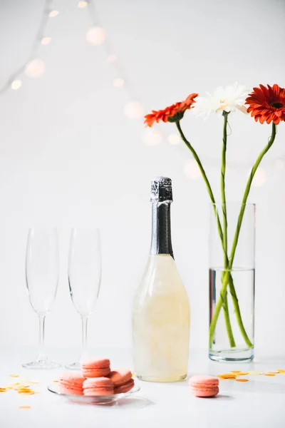 Vista de cerca de macarrones, copas vacías, botella de champán y ramo de flores de gerberas sobre fondo gris - foto de stock