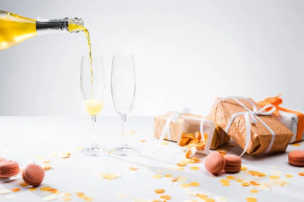 Close up view of pouring yellow champagne into glasses process, macarons and arranged gifts on grey backdrop — Stock Photo