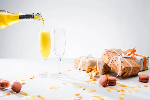 Vista de cerca de verter champán amarillo en el proceso de vasos, macarrones y regalos arreglados sobre fondo gris - foto de stock