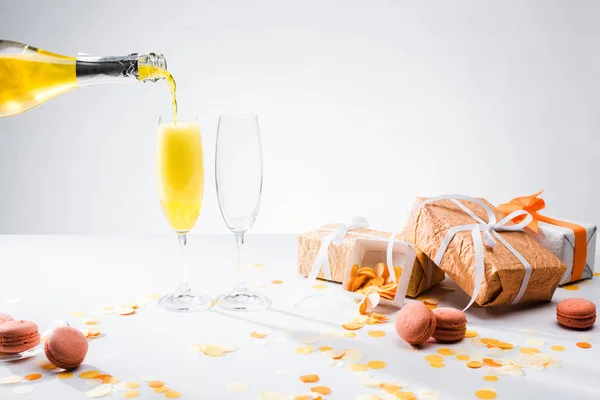 Vue rapprochée de verser du champagne jaune dans le processus de verres, macarons et cadeaux arrangés sur fond gris — Photo de stock