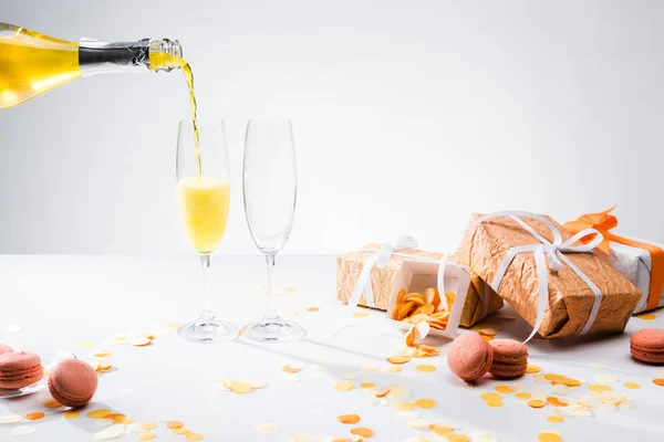 Vista de cerca de verter champán amarillo en el proceso de vasos, macarrones y regalos arreglados sobre fondo gris - foto de stock