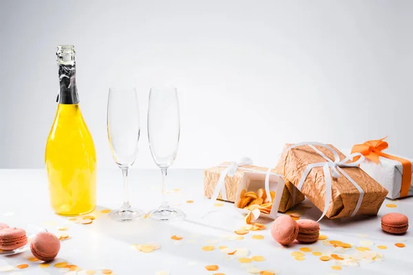 Vista da vicino della bottiglia di champagne giallo, bicchieri vuoti, macaron e regali disposti su sfondo grigio — Foto stock