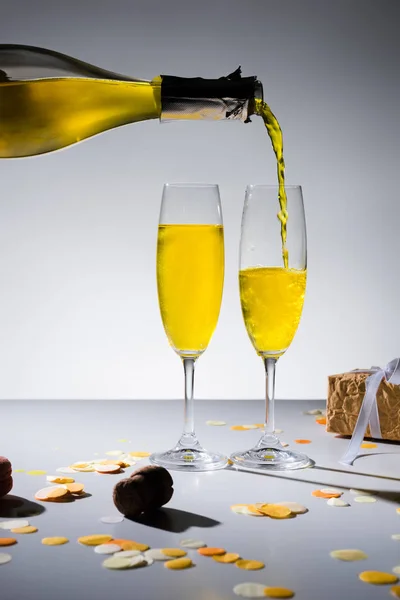 Vue rapprochée de verser du champagne jaune dans le processus de verres et cadeau enveloppé sur fond gris — Photo de stock