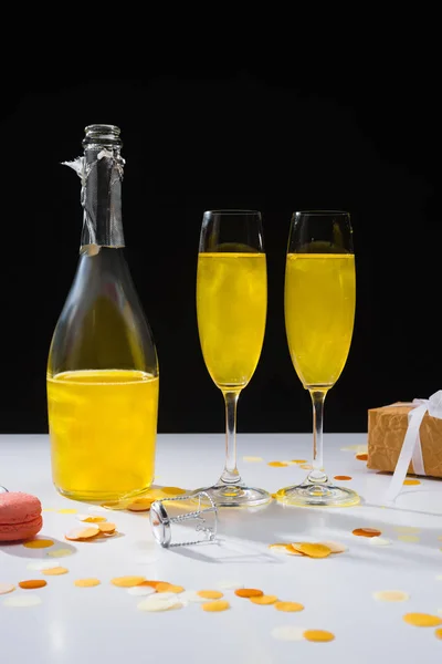 Vista da vicino di bottiglia e bicchieri di champagne giallo, macaron e regalo sulla superficie su sfondo nero — Foto stock