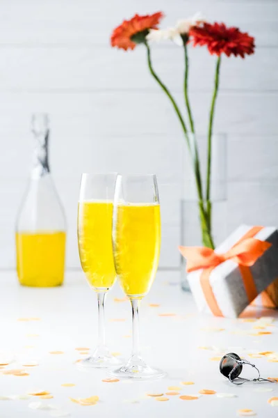 Selective focus of glasses of yellow champagne, bottle and gerbera flowers in vase on grey background — Stock Photo