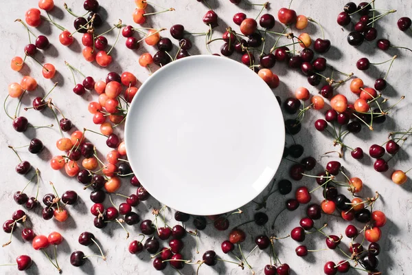 Сверху вид на пустую круглую белую тарелку и спелые сладкие вишни на белом — стоковое фото