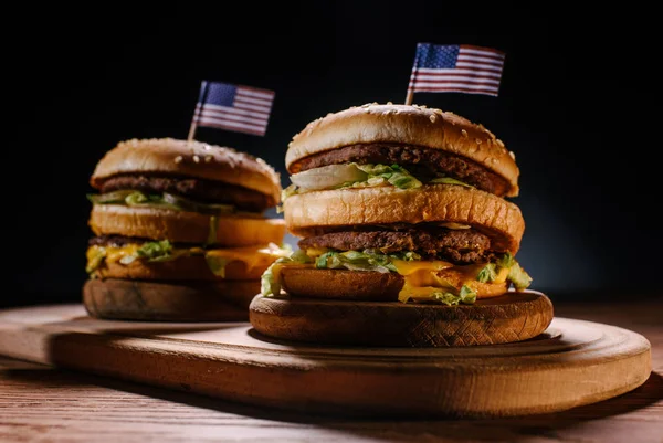 Close-up de deliciosos hambúrgueres com pinos de bandeira dos EUA em tábua de corte de madeira em preto — Fotografia de Stock