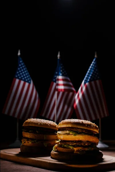 Sabrosas hamburguesas en tabla de cortar de madera frente a banderas de estados unidos pequeños - foto de stock