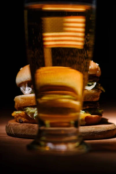 Vista sobre hambúrguer saboroso na placa de corte de madeira através de vidro de cerveja — Fotografia de Stock