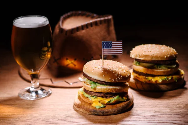 Sabrosas hamburguesas en mesa de madera con sombrero de vaquero americano y vaso frío de cerveza - foto de stock