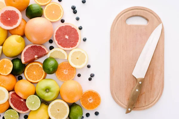 Top view of citrus fruits with blueberries and wooden cutting board with knife on white surface — Stock Photo