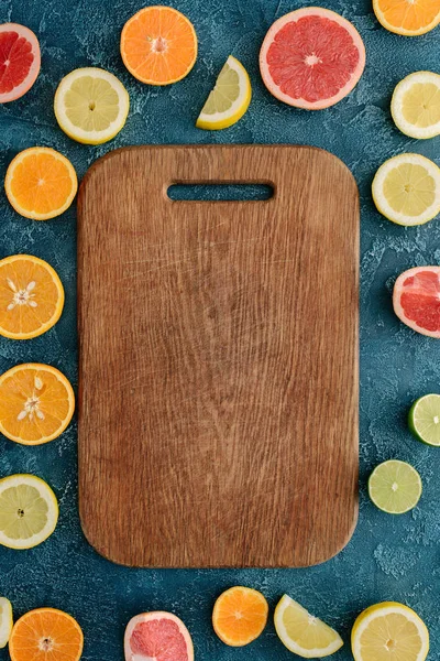 Top view of wooden cutting board surrounded with citrus fruits slices on blue concrete surface — Stock Photo